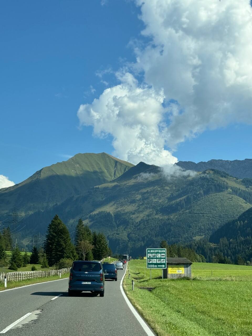 A row of vehicles drive down the road next to bright green grass with a mountainous landscape in the background.